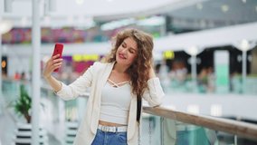 Young woman with curly hair wearing stylish clothes standing on mall terrace, looking at camera on smartphone and recording selfie video, blurred background. Blogger streaming online