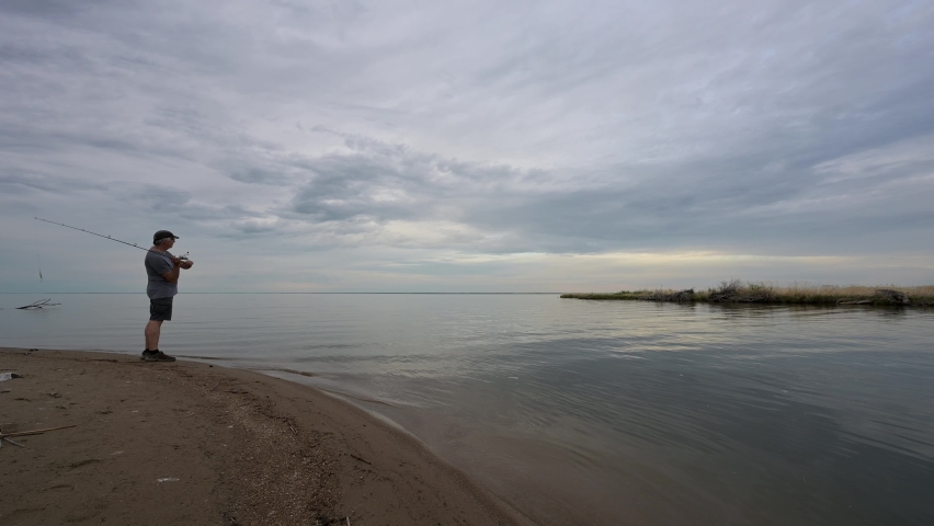 Man standing on beach fishing. He casts into the water. Sky is filed with gray clouds.
