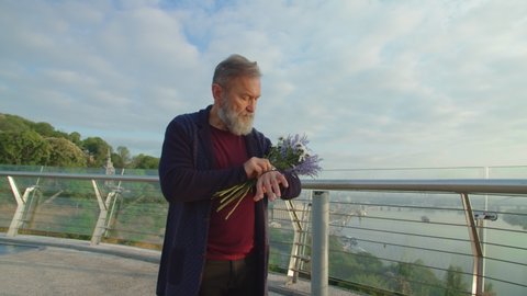 Senior bearded man with bouquet of flowers waiting for spouse, checking time on hand watch. Midsection of loving old husband waiting for wife outdoors. Romance and eternal love concept