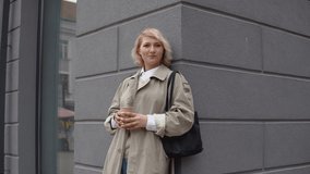 Young blonde business woman in a beige trench coat and white blouse with a black bag and takeaway coffee in her hands stands leaning against the wall of the building