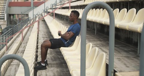 Zoom in view of tired black sportsman sitting on plastic seat and relaxing during break in training on stadium