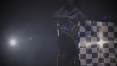 Side view of silhouette young woman waving checkered race flag to signal the start of racing event. Brunette posing in dark smoky studio with backlight. Close up. Slow motion ready, 4K at 59.94fps.