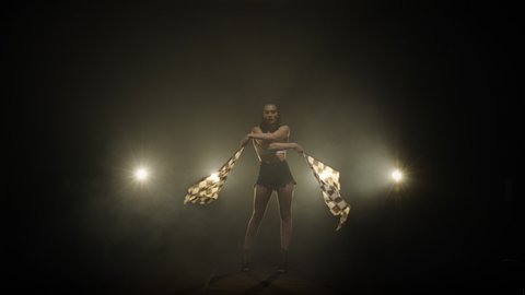 Pretty young woman waving checkered race flags while performing in racing competition. Silhouette of brunette poses full length in a dark smoky studio with backlight. Slow motion ready, 