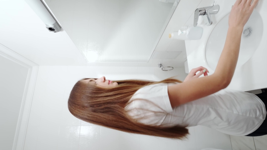Side view of smiling young woman in white shirt and with beautiful long hair looking at mirror and doing makeup in bathroom. Concept of process makeup after shower at home or hotel.
