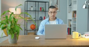 Confident hardworking asian man sitting at the table and working on computer in specially designeted place at home, front view