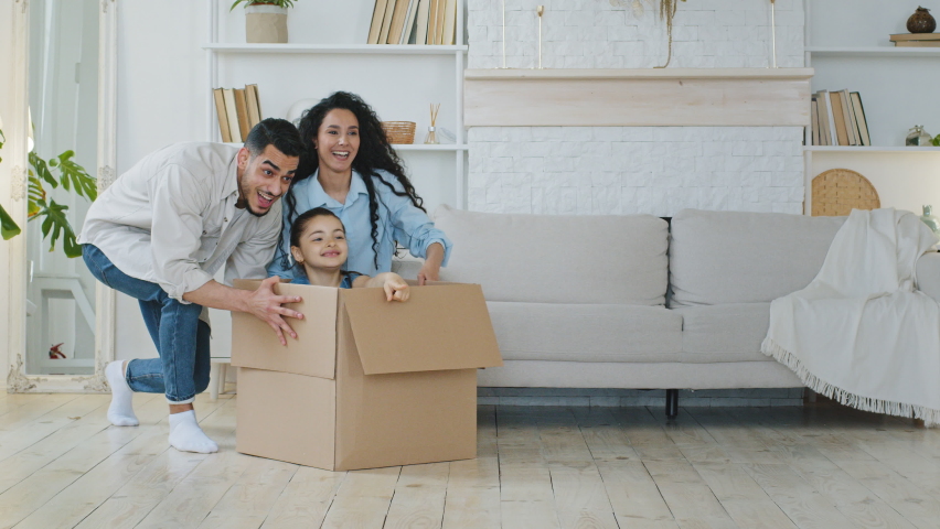 Funny active multiracial parents mom dad multiethnic family playing on moving day at new home pushing cardboard box with cute little child girl daughter kid sit inside having fun packing relocate Royalty-Free Stock Footage #1073673098