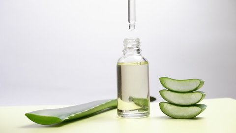 A drop of natural juice flows from a cut aloe leaf into a green jar with aloe vera gel on a white background.