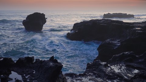 A tidal wave splashing on the rock at sunset hour. Raging sea waves hitting stones. Water element in its fierce power at blue hour in tropical paradise. Balinese ocean waves and black sharp reef.