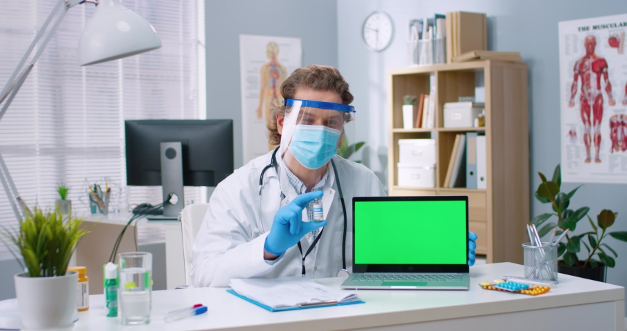 Doctor in protective medical mask and gloves looks into camera and talks about vaccine holding test tube in his hands with chromakey laptop. Royalty-Free Stock Footage #1073686316