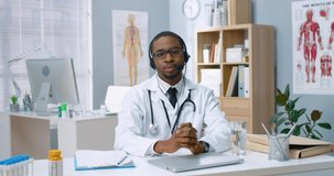Portrait of young African American doctor in glasses and headphones with microphone sitting in hospital office and recording video message to patients talking about symptoms of virus.