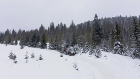 A small tractor removes white snow from a snow-covered road up a hill so that cars can drive through evergreen forests, aerial UHD 4K video