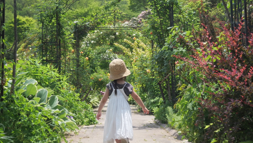 Little girl in a straw hat is walking in a beautiful green plant garden with rose flowers. 3 years old preschool toddler child back view. Early summer season. Point of view shot of Gimbal motion Royalty-Free Stock Footage #1073697728