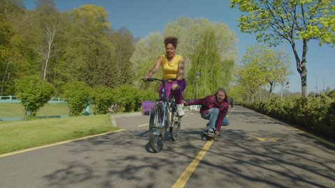 African female cyclist towing hipster friend on longboard skateboard while cheerful active beautiful diverse multiracial women enjoying recreation together, cycling and skateboarding in public park.