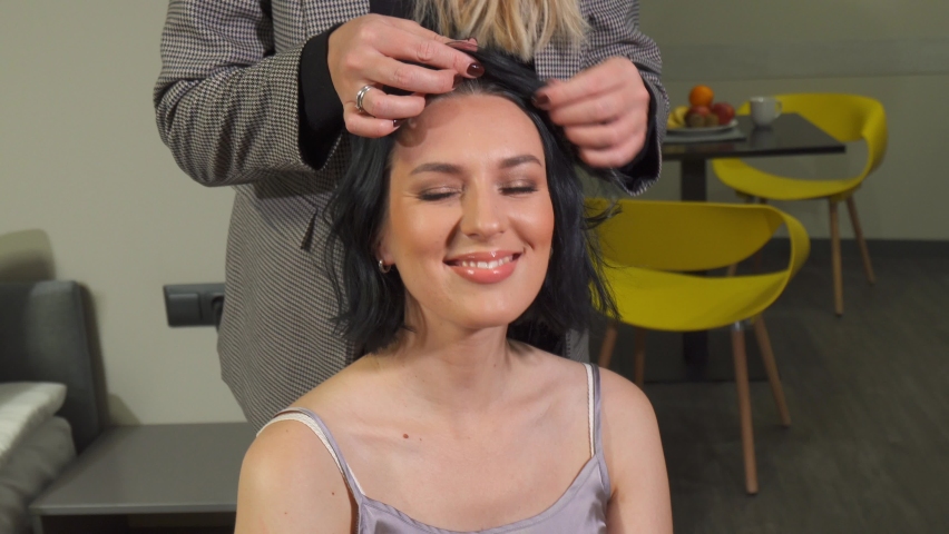 a beautiful brunet girl with shoulder-length hair, the hairdresser straightens her curls with his hands. the girl is satisfied, smiles and dances slightly while sitting still