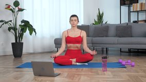 Beautiful fit girl athlete sits meditating in lotus position with closed eyes on sport fitness blue mat, breathes deeply. Calm sportive young woman in sportswear practices yoga at home in living room