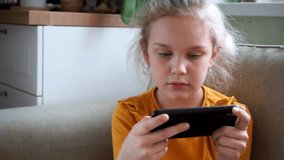 7-year-old girl in a yellow longsleeve is enthusiastically playing a game on her smartphone, discussing the action by ear. she is sitting sitting on the couch.