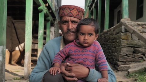 close up shot of an Indian traditional Himachali old man sitting and holding a little kid in his hand with wooden structured houses in the background looking at the camera