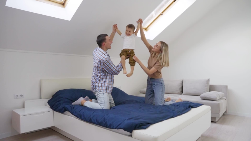 Happy young family with boy child playing enjoying time together at home jumping on bed in bedroom, mom and dad holding boy, laughing having fun. Slow motion.