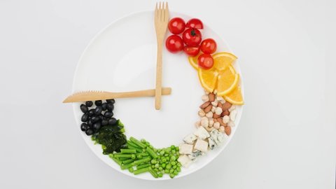 Colorful food and cutlery arranged in the form of a clock on a plate. Woman's hand takes an olive. Intermittent fasting, diet, weight loss, lunch time concept.