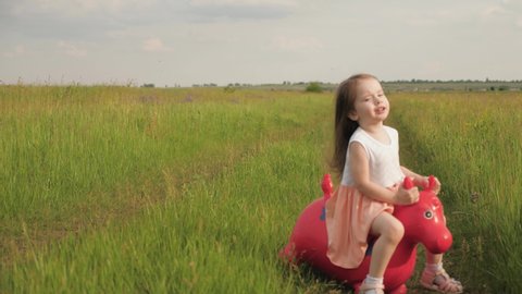 Little girl, daughter playing in field outdoors. Family and childhood, vacations. Happy little girl is playing in park, jumping on an inflatable toy horse. Kid plays on playground in summer in nature.