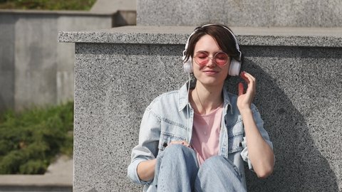 Young calm happy woman 20s wearing denim shirt pink t-shirt glasses listen music in headphones rest relax in city sit on concrete stairs sunshine day outdoors nature. Urban lifestyle sun light concept