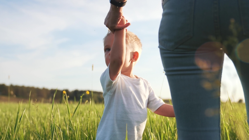 Happy family. Mom holds her kid son by hand in park. Mother and son are walking through park. Kid holds his mother by the hand in the green grass. Happy family concept. Happy kid in the park with mom. Royalty-Free Stock Footage #1073717510