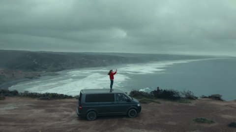Adventurous woman in red sweater stand on roof of her camper van, make video for social media on smartphone. Aerial shot of travel blogger in epic cinematic location on ocean cliffs.Wanderlust concept