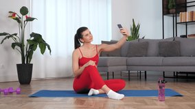 Positive fit sports girl records stories after workout exercise training at home. Smiling sportive woman fitness blogger sitting on yoga mat listen tells online video call on selfie camera smartphone