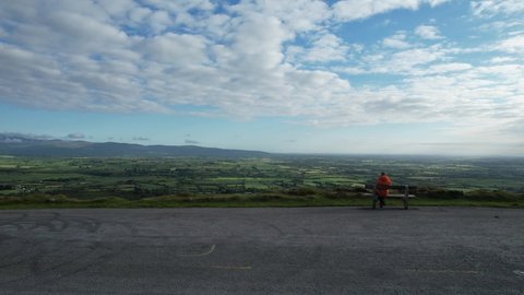 A man sits on a bench near the edge of the road admiring the views at Vee Pass, a v-shaped turn on the road leading to a gap in the Knockmealdown mountains in Clogheen county Tipperary, Ireland Adlı Stok Video