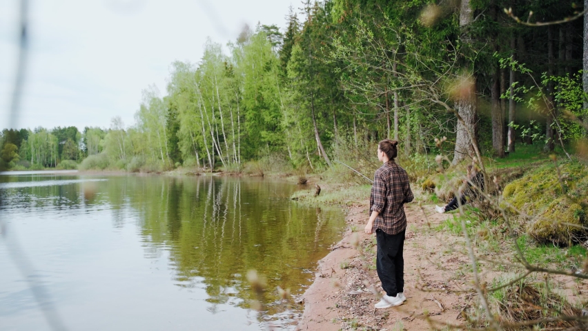 A beautiful young woman in a checkered shirt is fishing on the lake. A woman with a fishing rod stands on the bank of the river in the forest.
