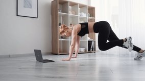 Fitness training online. Home gym. Physical exercise. Athletic woman in sportswear doing running plank with web video tutorial on laptop on floor at light room interior.