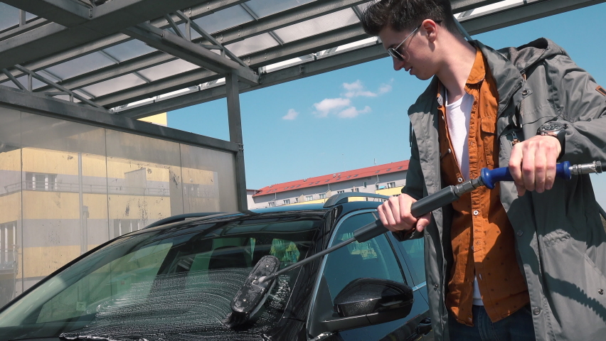 Cleaning dirt after trip man with glasses washes car hands. He smears soap all over car. Use water jet to wash dust.