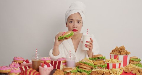 Hungry woman bites delicious hamburger drinks fizzy drink consumes fast food wears red lipstick domestic robe has bad eating habits isolated over white background breaks diet. Cheat meal concept