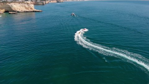 Aerial, drone shot, following a boat, dragging people on a inflatable toy, on the coast of Algarve, on a sunny day, in Carvoeiro, Portugal