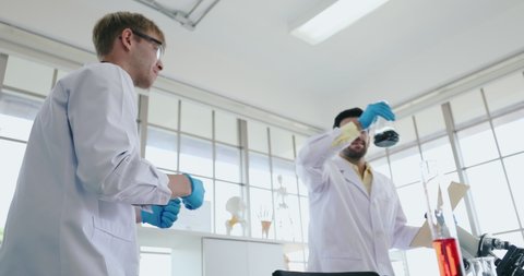 two young funny male scientists wearing white lab coat dance around in the science lab holding glass flask with chemical inside. Concept of crazy mad scientist lab experimentation.