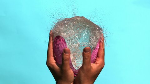 Hands Holding Pink Water Balloon and Bursting it in Slow Motion on a Blue Background