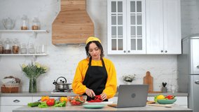 Woman housewife in apron slices bell pepper on cutting board, tells teacher chef study online video call conference webcam chat laptop computer, distance education culinary course in home kitchen