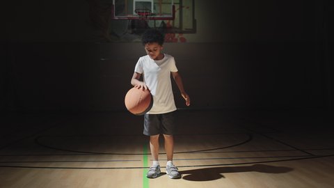 Slowmo shot of cute boy standing in spotlight on dark indoor basketball court and dribbling ball