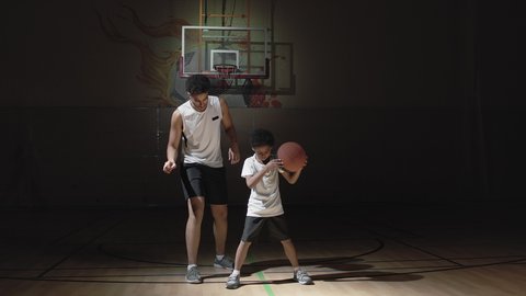Handheld tracking shot of young male basketball player in sportswear teaching his little son how to dribble ball on dark indoor court