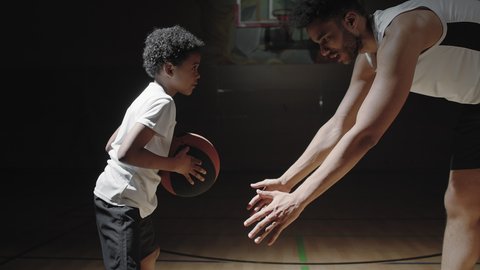 Handheld shot of black male basketball player talking and teaching boy how to dribble ball on dark indoor court
