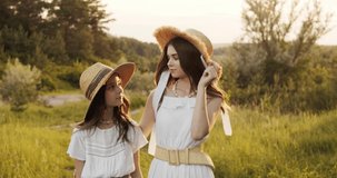 Two girl teenagers in white dresses and straw hats with flowers bouquet posing on nature landscape at sunny summer day