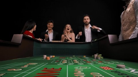 Ggroup of men and women stand at green craps table in casino. Charming lady rolls dice and wins. Company rejoices with delight and celebrates the victory. View from inside of game table. Slow motion.
