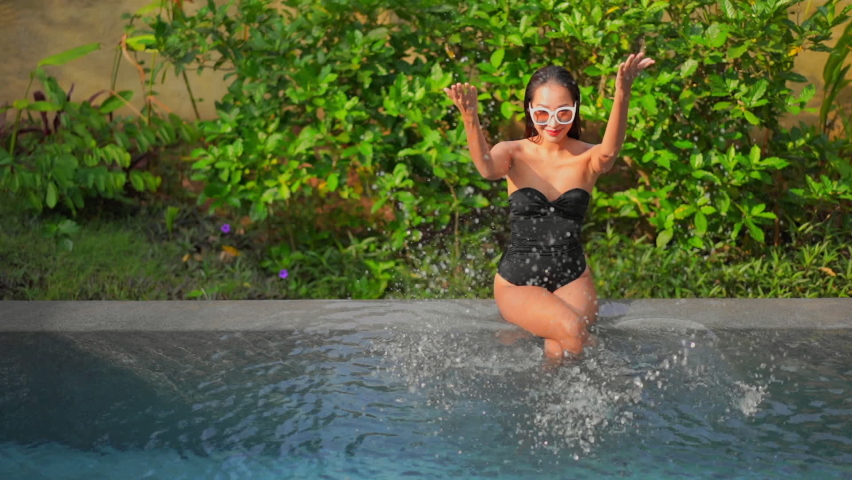 A beautiful fit Asian woman in a black bathing suit and sunglasses sits on the edge of a resort pool with feet in the water together. Happy girl relaxing splashing water with both hands