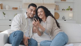 Hispanic family spaniard man and ethnic beautiful curly woman multiracial couple sitting on sofa at home living room girl holding mobile phone looking together in smartphone screen talking surprised - Powered by Shutterstock - Get 15% off with code: PIKWIZARD15