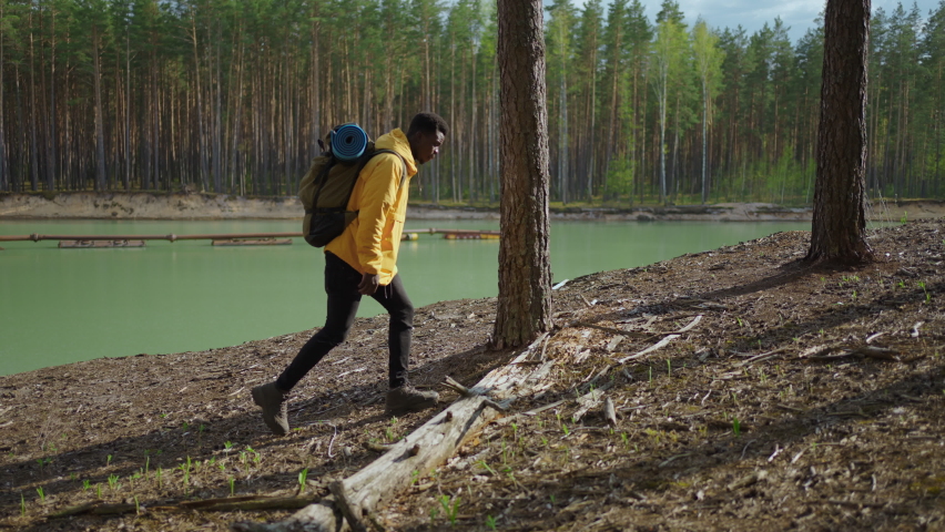 A young black man in a yellow cursor climbs up with a backpack uphill in the woods looking at the lake. Slow motion