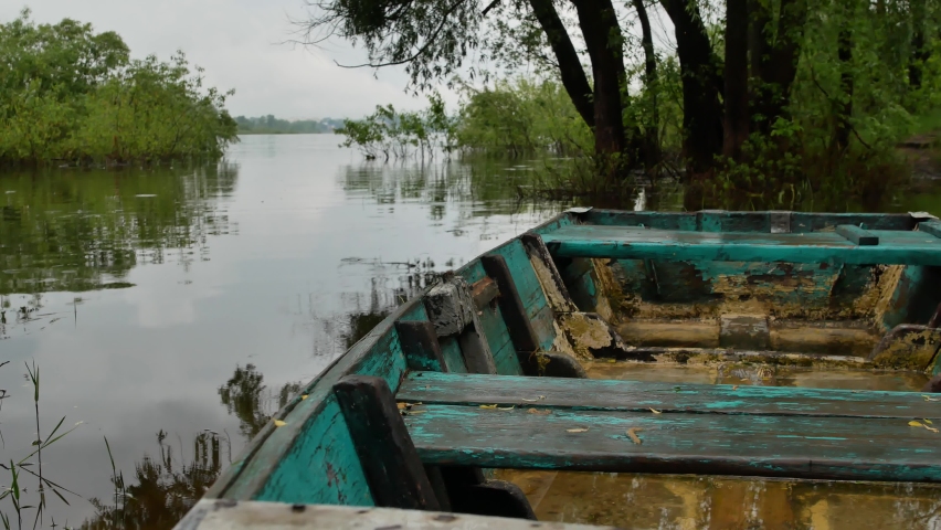 4K summer video background. An old wooden small fishing boat moored by the river on a rainy summer or spring day against the backdrop of a large river and green trees.
