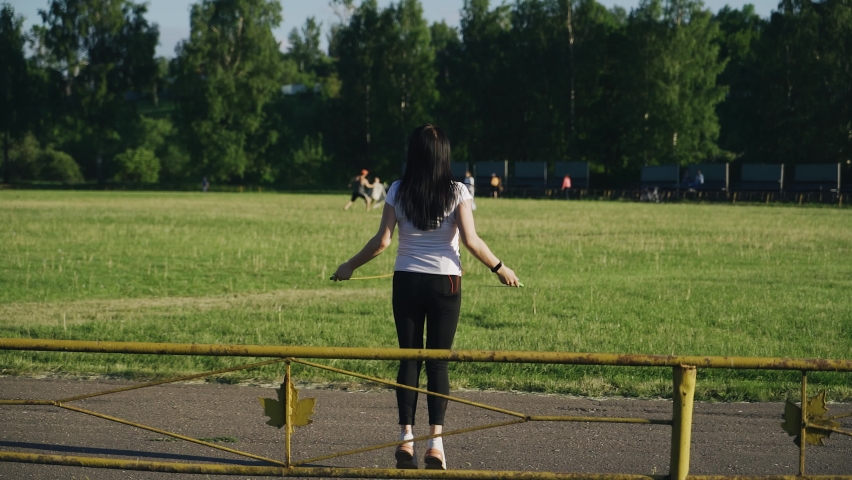 A girl in black leggings and a white t-shirt jumps over a jump rope at the stadium. She does fitness classes