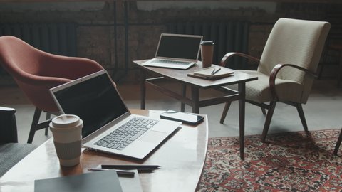 Laptops, coffee cups, notebooks and other stationery on work desks in modern loft coworking space with stylish chairs and furniture