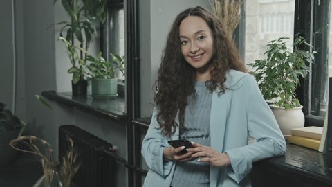 Medium slowmo portrait of young attractive Mixed-race woman with long curly hair smiling to camera while standing at windowsill in modern office using smartphone