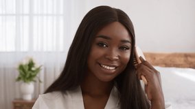 Cheerful Black Female Brushing Long Hair Smiling To Camera Sitting In Bedroom Indoors. African American Female Enjoying Haircare Routine Combing Hair With Hairbrush At Home